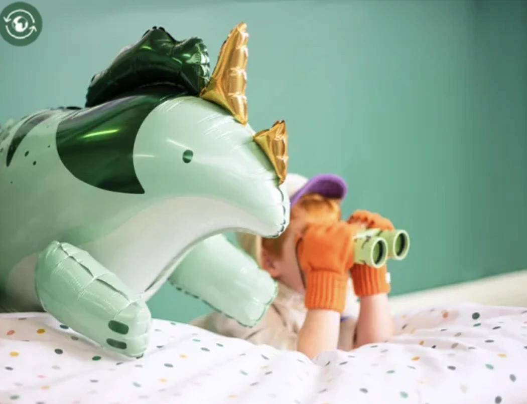 A triceratops foil balloon next to a child exploring with binoculars.